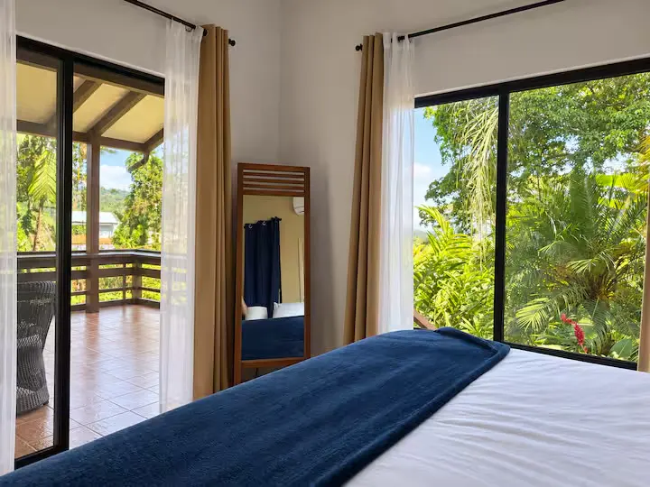White bed with a navy throw beside tropical greenery and a balcony